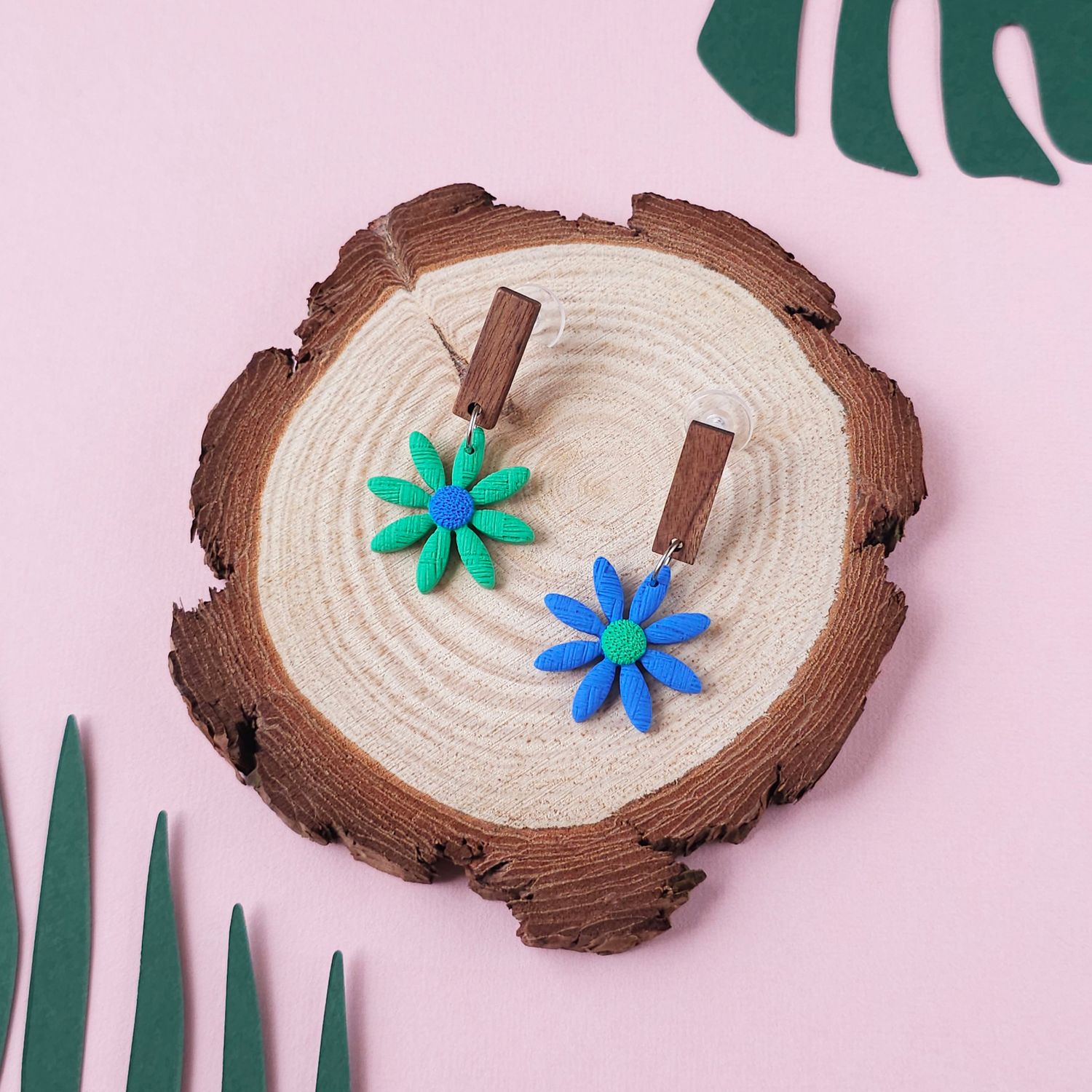 A pair of daisy earrings, one green with a blue centre and one blue with a green centre, with a dark wood rectangular top.