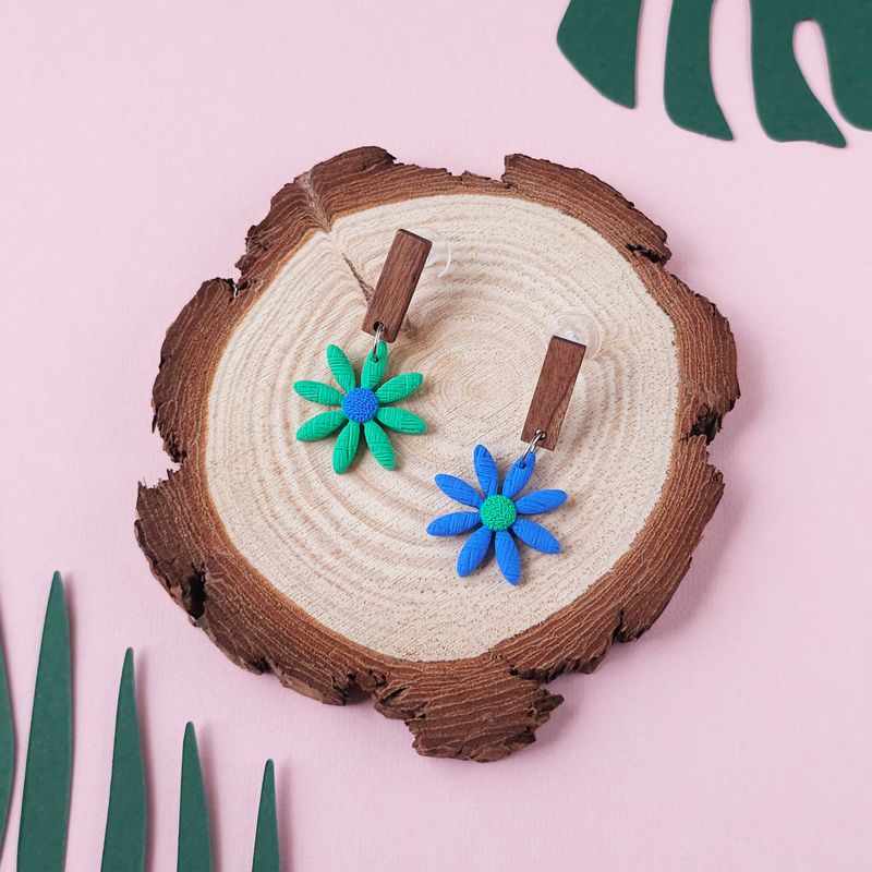 A pair of daisy earrings, one green with a blue centre and one blue with a green centre, with a dark wood rectangular top.