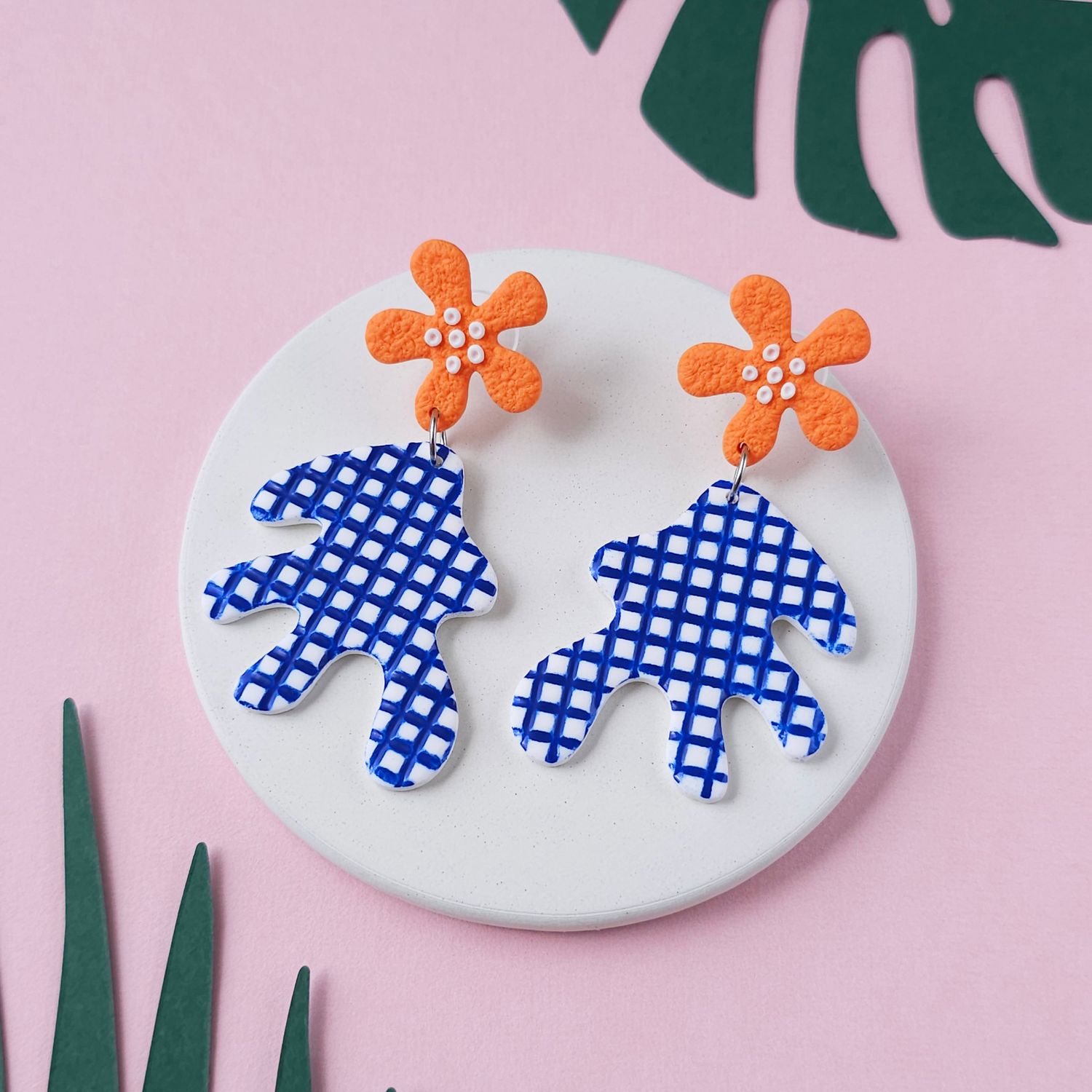A pair of earrings that features orange flower-shaped studs with white dot details and checkered, abstract shapes below.