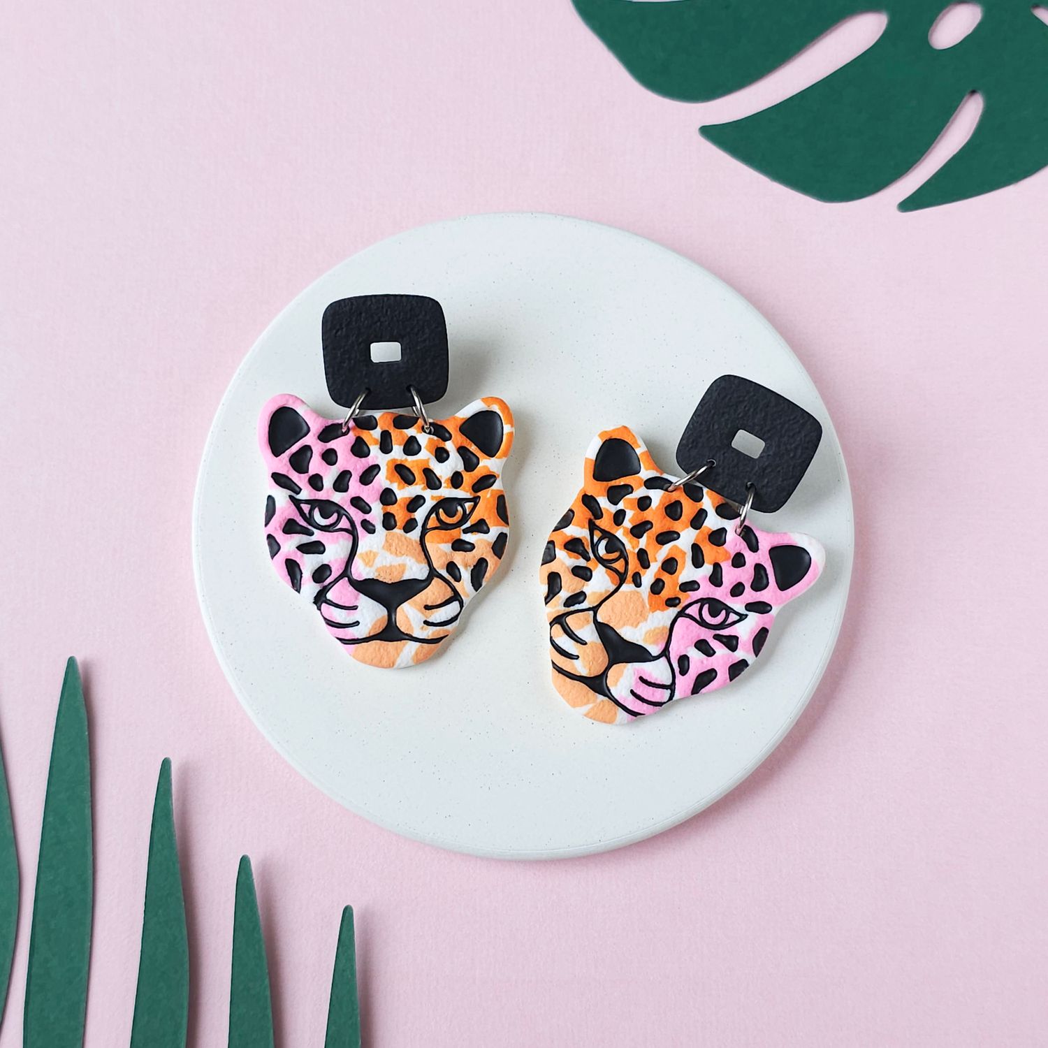 A pair of statement earrings that have a black square stud and a jaguar head design with orange, pink, and black spots.