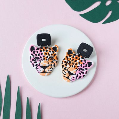 A pair of statement earrings that have a black square stud and a jaguar head design with orange, pink, and black spots.