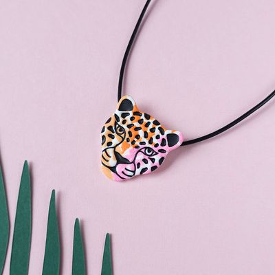 A necklace with a jaguar head pendant in orange, pink, black, and white. The pendant hangs from a black leather cord.