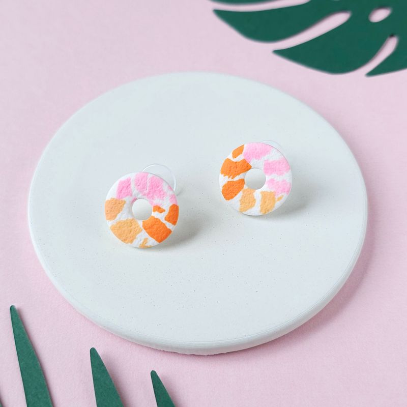 A pair of small donut-shaped stud earrings with a white base and a pattern of pink and orange irregular shapes.