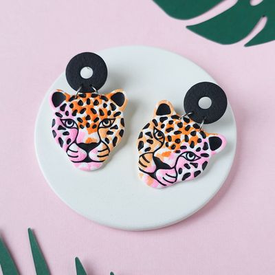 A pair of jaguar face earrings that have a black circular top, with a leopard face design in orange, pink, and black.