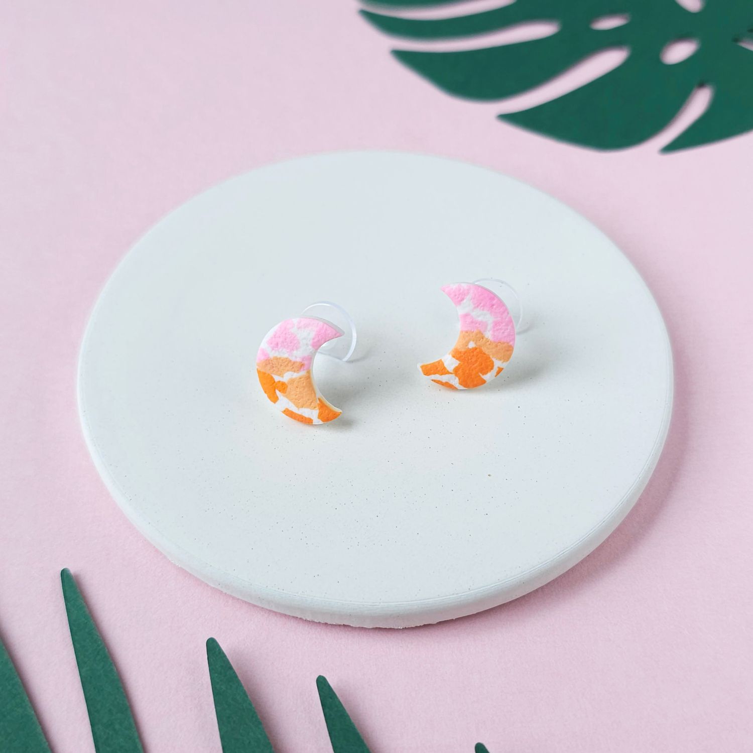 A pair of crescent moon stud earrings. The earrings have a speckled design in pink, orange, and white.