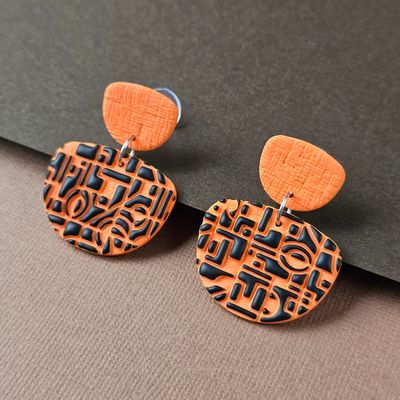 The earrings feature a textured orange top piece and a larger, rounded bottom piece with a black geometric pattern.
