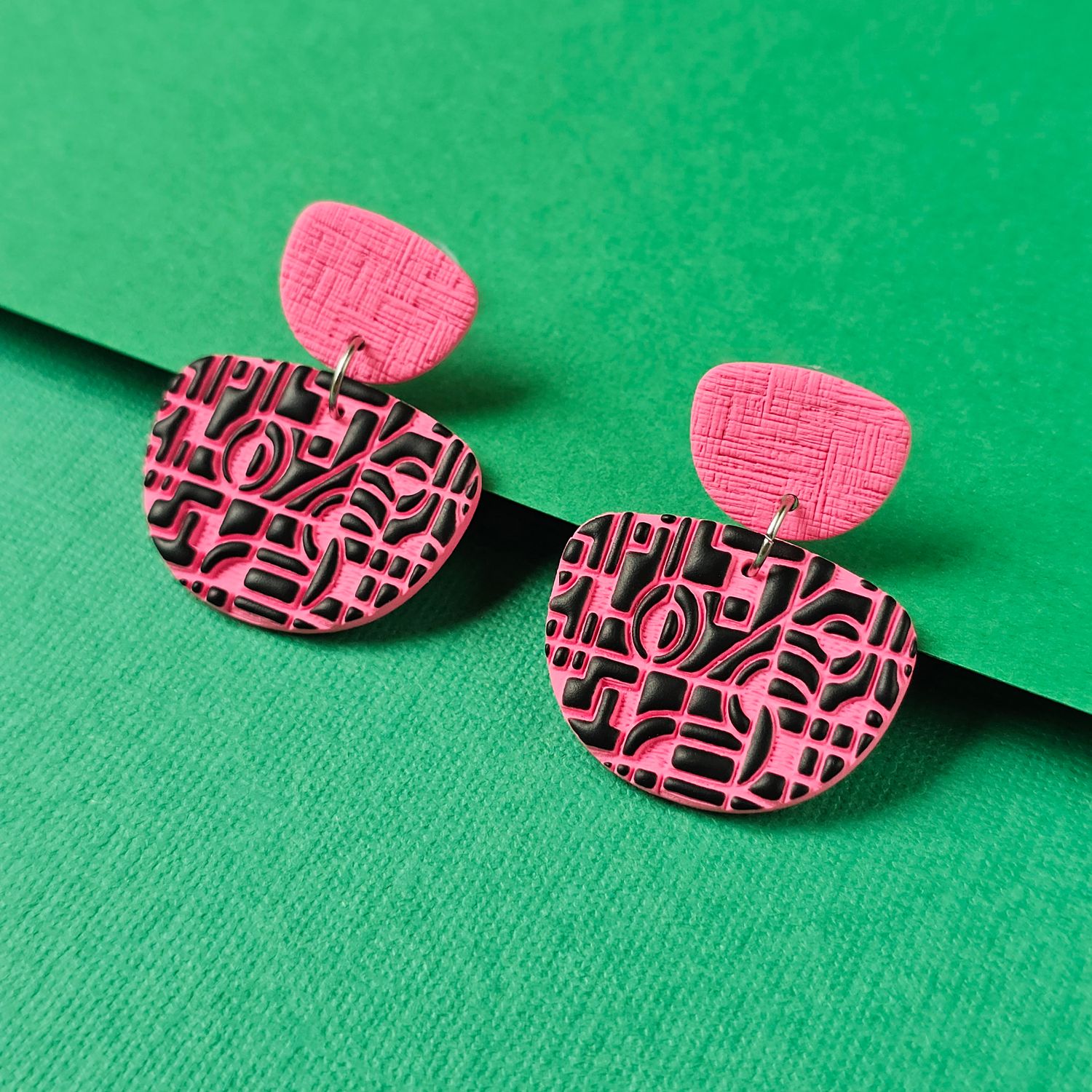 A pair of pink and black earrings. The main part of the earrings is a pink, rounded square with a black geometric design.