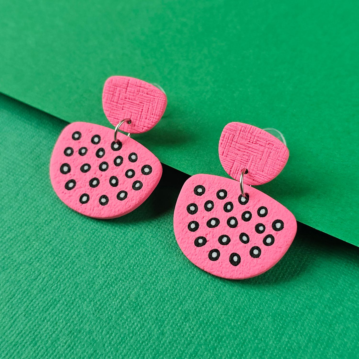 A pair of pink earrings, with a textured, oval top and a larger, irregular shape below, decorated with black circles