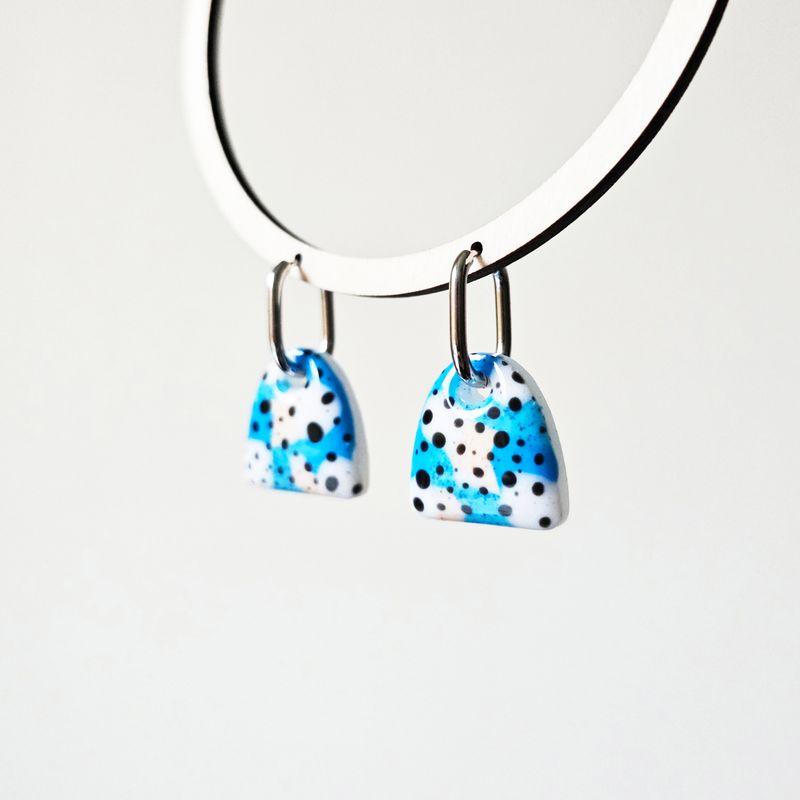 A pair of abstract dangle earrings. Each earring has a resin charm with white, blue and black splatter designs.