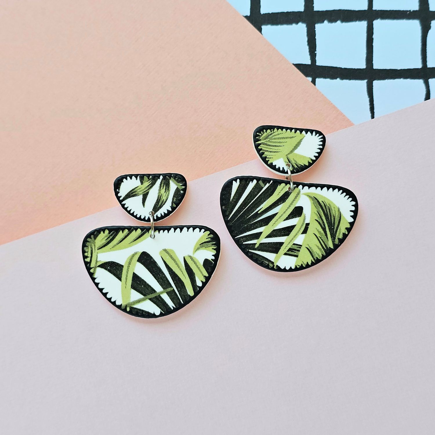 A picture showing a large pair of earrings with a green foliage print and black zigzag edges.