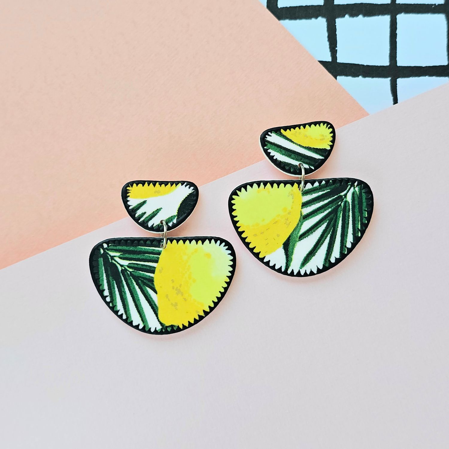 A picture showing a large pair of earrings with green foliage and a side of a lemon print on them, and a black zig zag edge