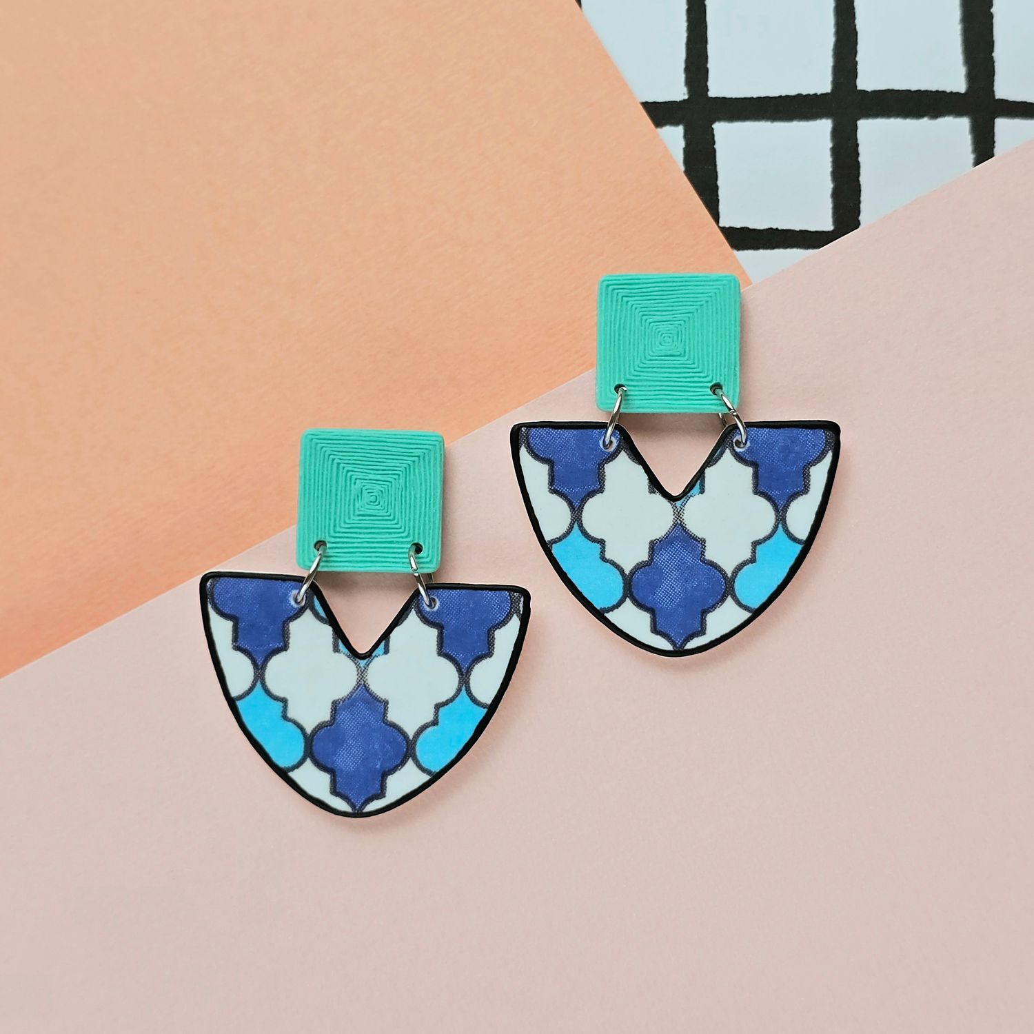 A picture showing a large pair of earrings with a geometric blue/white tile print and an aqua blue textured upper square bead