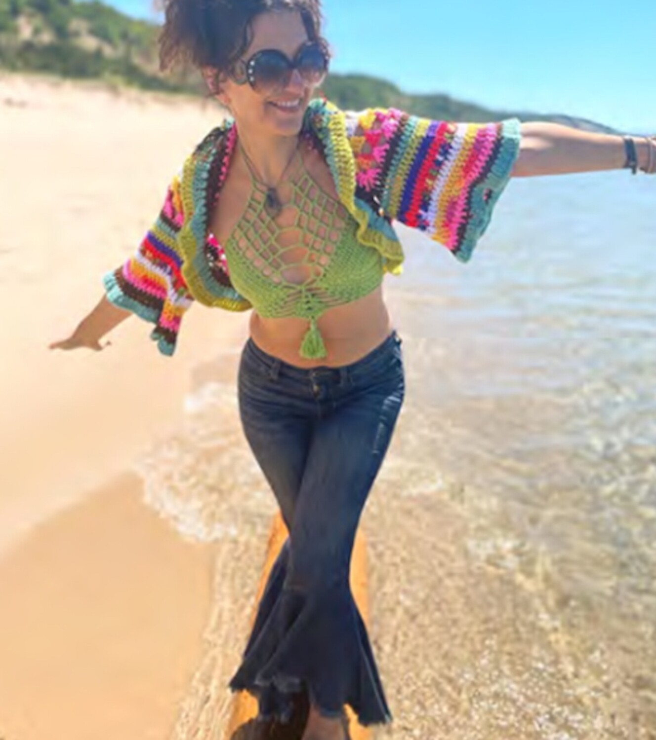 a light-skinned woman with her curly brown hair worn in a messy updo cavorts on a beach. Her arms are up and out "airplane-style". We see sand at her right, and turquoise water on her left with greenery in the distance. She's wearing jeans, a crocheted bikini top, and a multicolored crochet shrug, as well as large sunglasses and a chunky pendant. She's smiling and seems to be having fun!