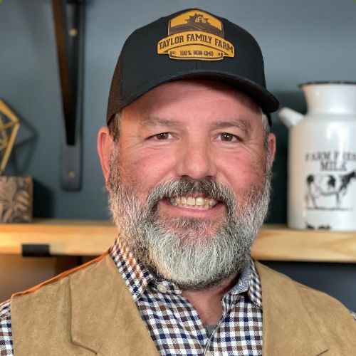 Farm Life with Farmer Johnny: Bearded man in a black "Taylor Family Farm" hat, plaid shirt, and tan vest, smiling.