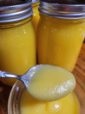 Golden yellow Ghee in glass jars with silver lids, with a spoonful of creamy Ghee over a clear bowl of Ghee.