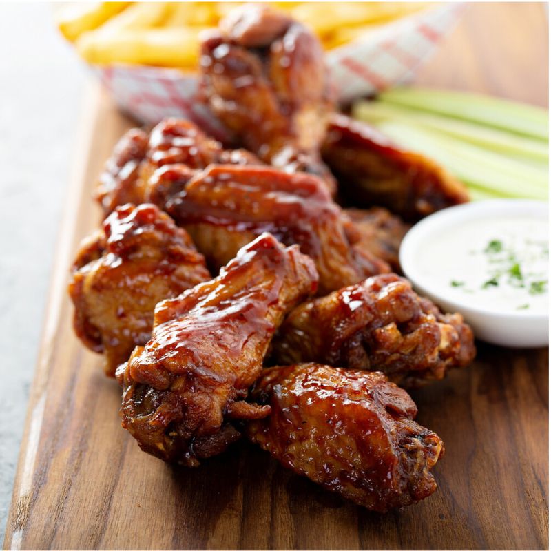 Wing Party Pack - 5 lb: Juicy, glistening brown BBQ chicken wings on a wooden board, with ranch dip, celery sticks, and fries