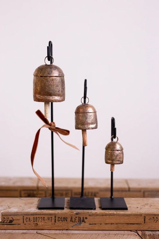 Three rustic metal bells hang from black iron stands of varying heights, creating a vintage, minimalist display