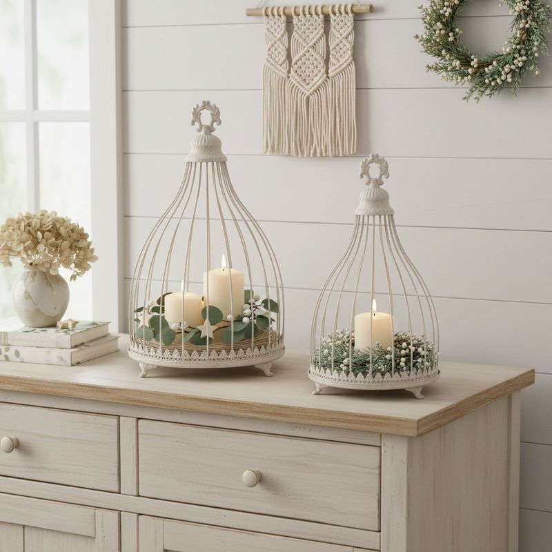 Two vintage style ivory metal caged cloches with ornate bases on feet decorated with candles and green foliage