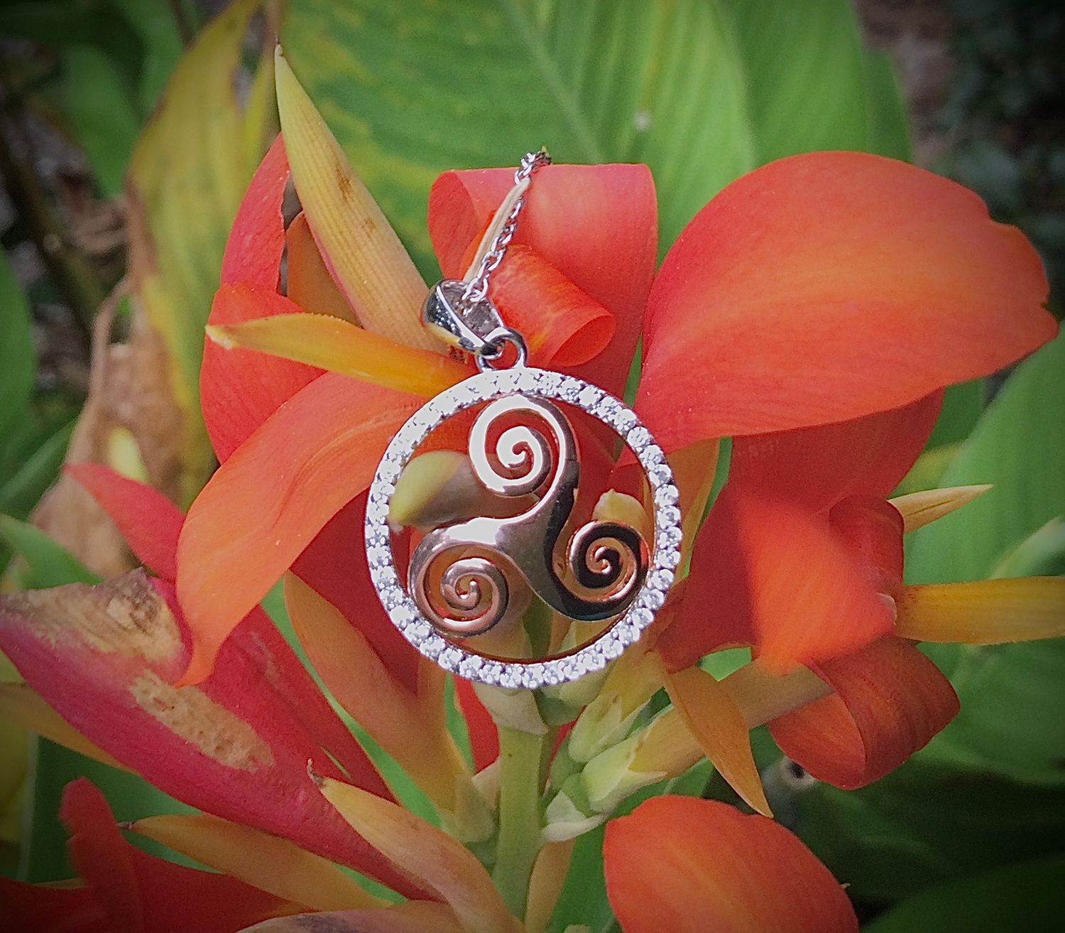 Stone Circled Rose Gold Triskele Sterling silver triskele pendant with rose gold plating, surrounded by sparkling cubic zirconia stones, shown on flower.