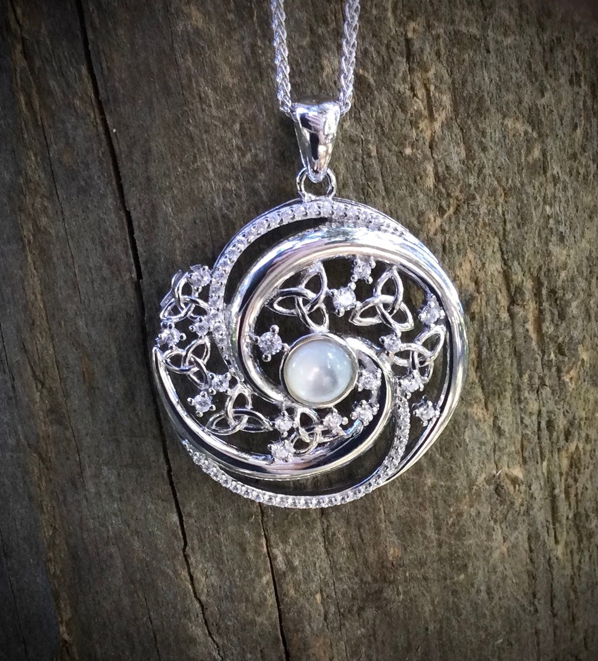 Arian Mother of Pearl Trinity Swirl Pendant Sterling silver Celtic pendant featuring a central Mother of Pearl stone, handcrafted with intricate knotwork, shown on bark.