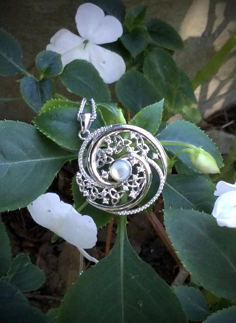 Irish-inspired sterling silver pendant with Celtic knot design and central Mother of Pearl gemstone, shown near plants. 
