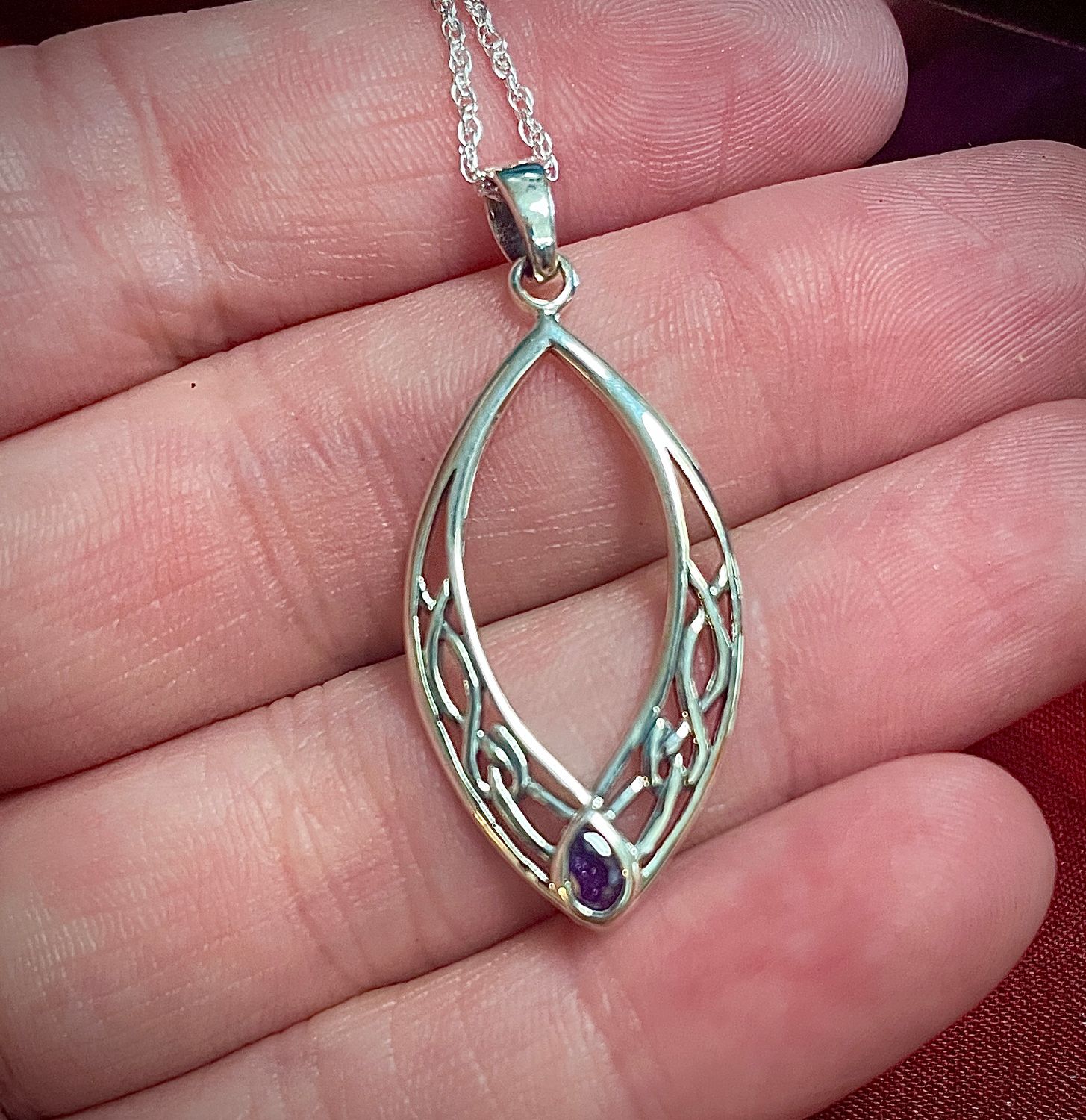 Oval-shaped Celtic knot pendant in sterling silver featuring a purple amethyst-style gem. Held in hand by model.