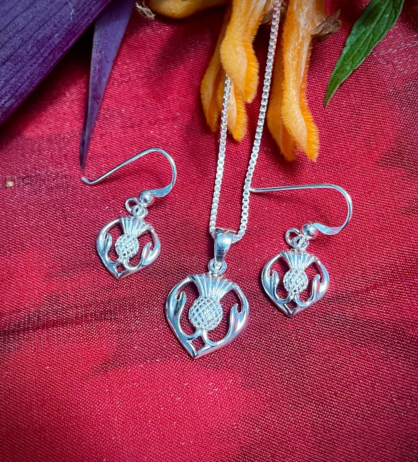 Sterling Silver Scottish thistle pendant and drop earrings jewelry set, highlighting Scottish heritage, shown on red fabric. 