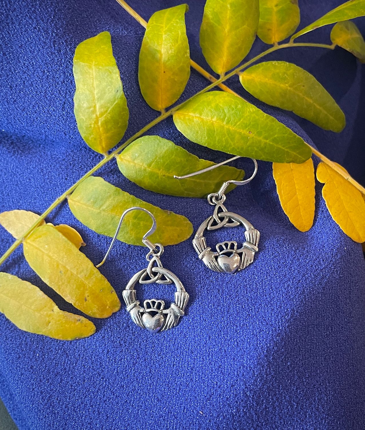 Elegant sterling silver Celtic Claddagh earrings with trinity knot, on blue fabric near autumn leaves.