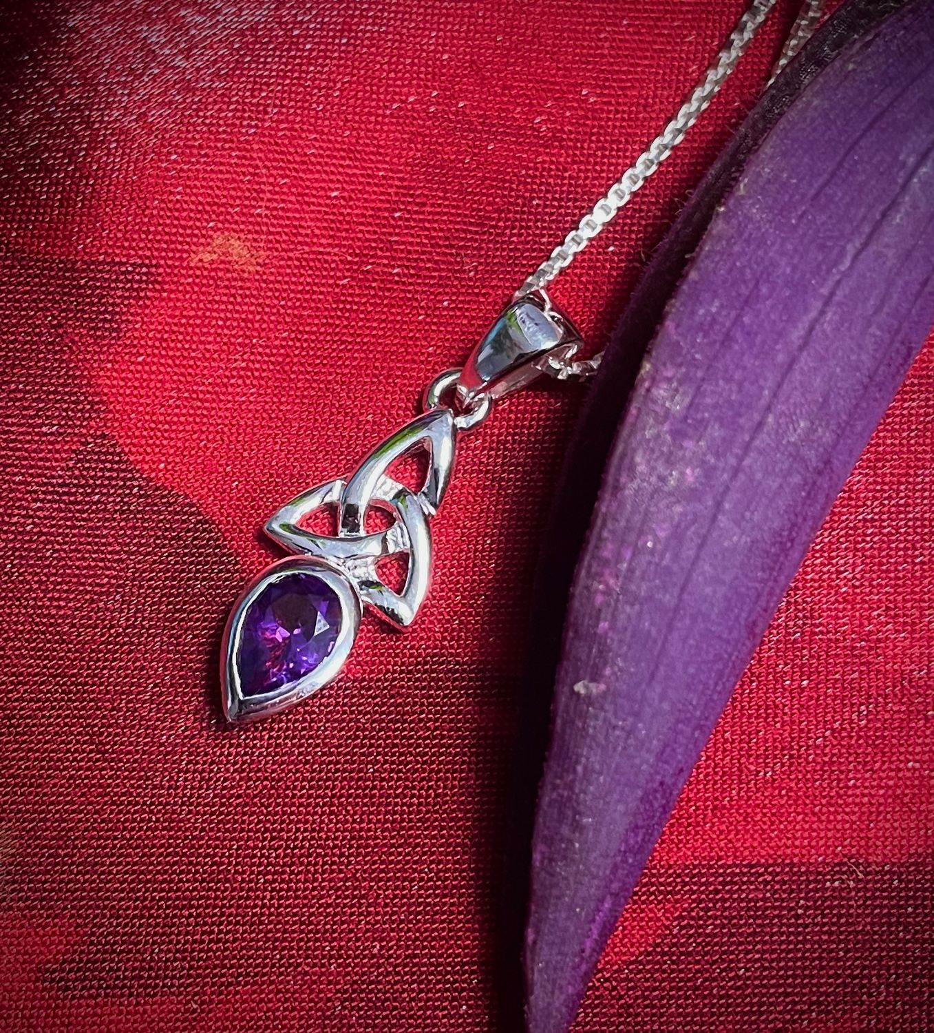 Sterling silver Trinity Knot pendant with amethyst February birthstone, symbolic Celtic jewelry, on red fabric. 
