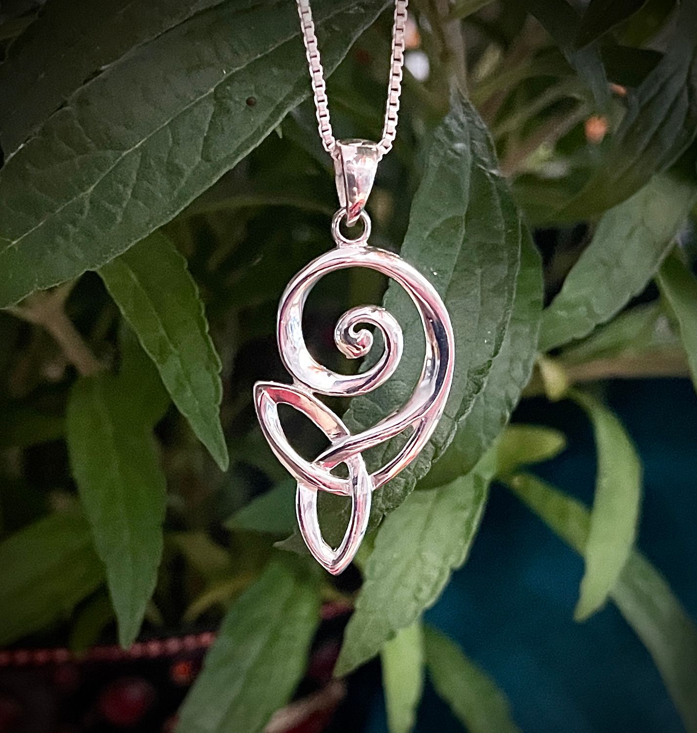 Sterling silver Trinity Knot Celtic pendant with swirl design, handcrafted heritage necklace, by green leaves.
