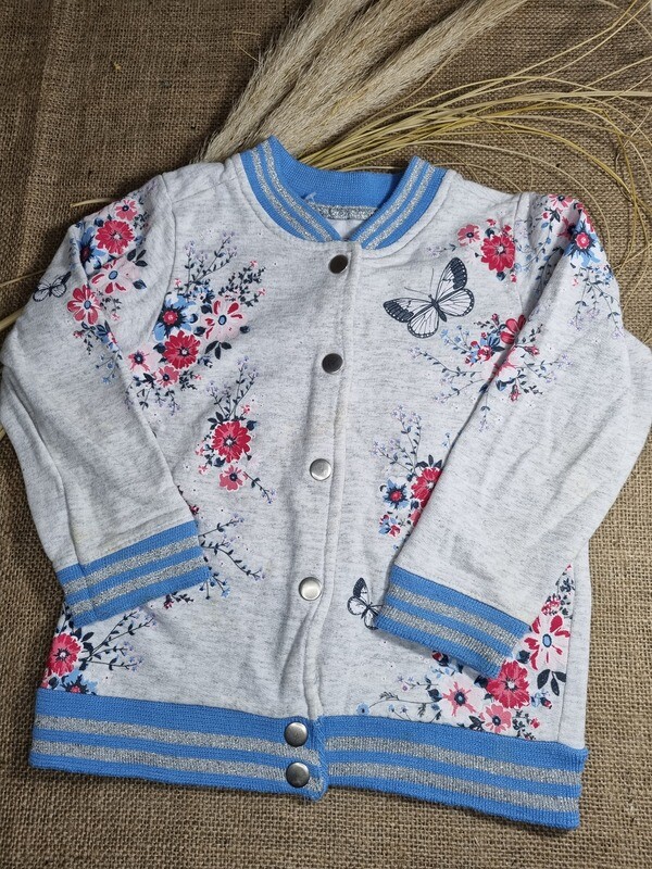 CAMPERA DEPORTIVA, COLOR GRIS ESTAMPADA CON FLORES Y MARIPOSAS, CUELLO, PUÑOS Y CINTURA ELASTIZADA EN CELESTE Y PLATEADO, MARCA POIM, TALLE 3. DETALLE DE MANCHITA ENTRE EL 2º Y 3º BROCHE.