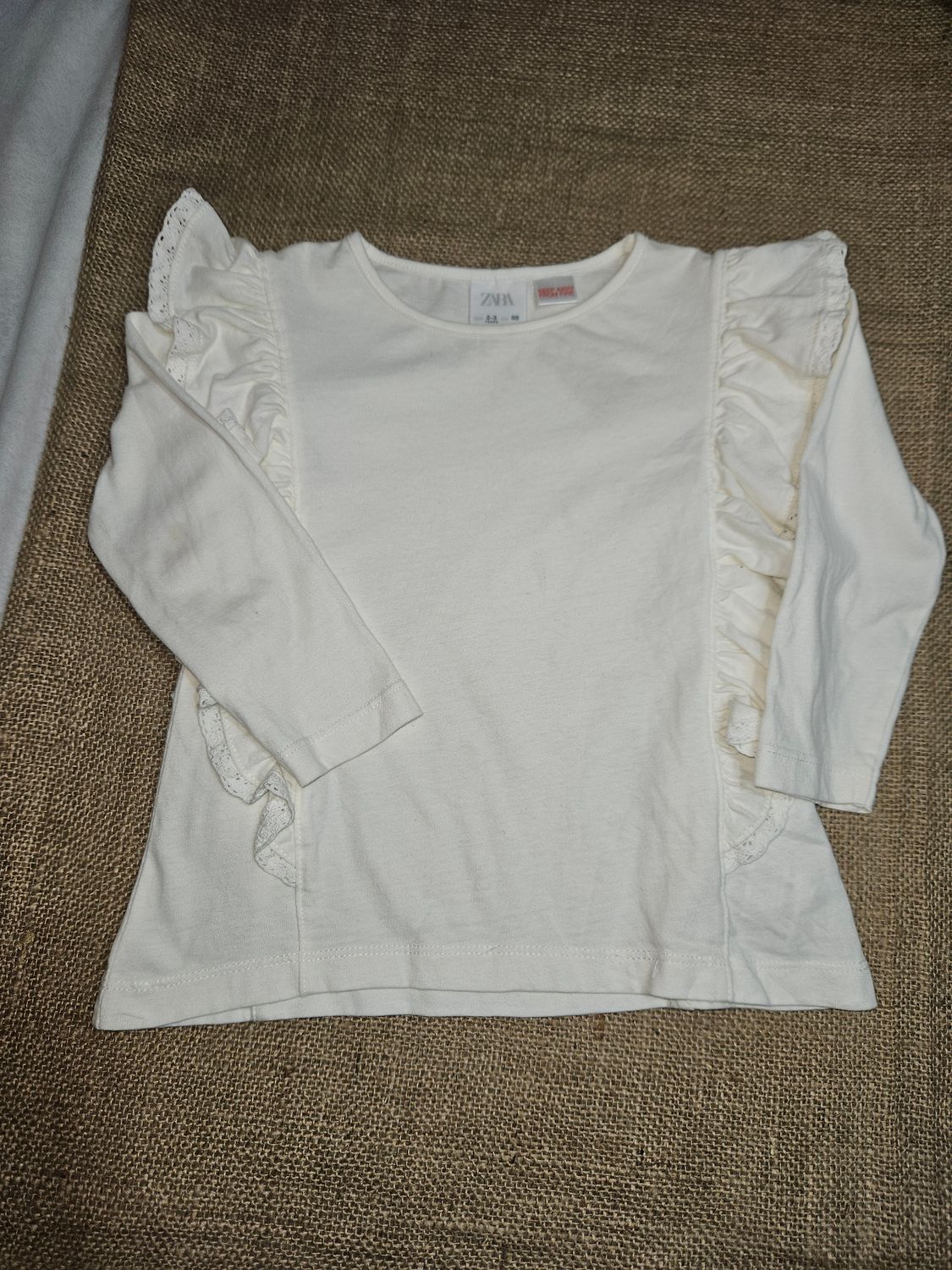 REMERA DE ALGODÓN, MANGA LARGA, CON VOLADOS. MARCA ZARA. TALLE 2-3. COLOR BLANCO. CON PEQUEÑO ROCE EN CUELLO