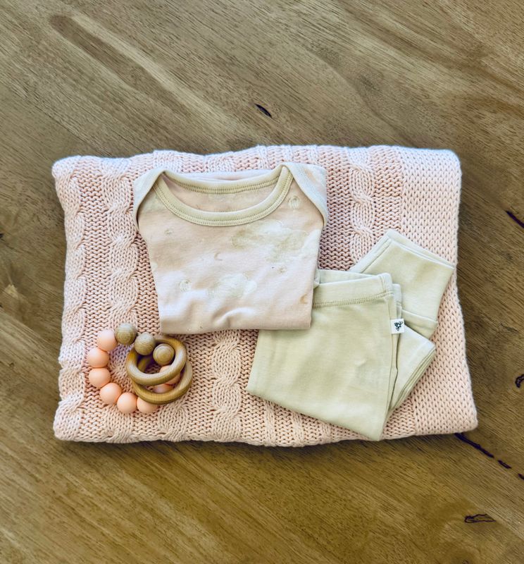 Neutral-toned baby girl gift set with Burt’s Bees onesies, pants, burp cloths, and a soft bear rattle on wooden table
