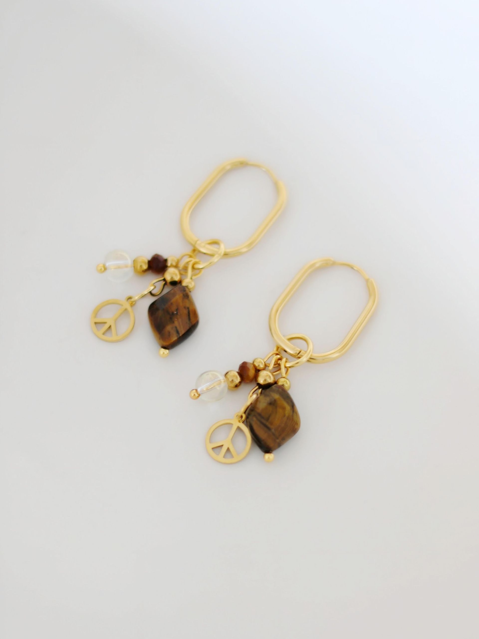 Ein Paar ovale Creolen in Gold mit langen Anhängern aus verschiedenen Perlen aus Tigerauge, Edelstahl und Peace-Zeichen