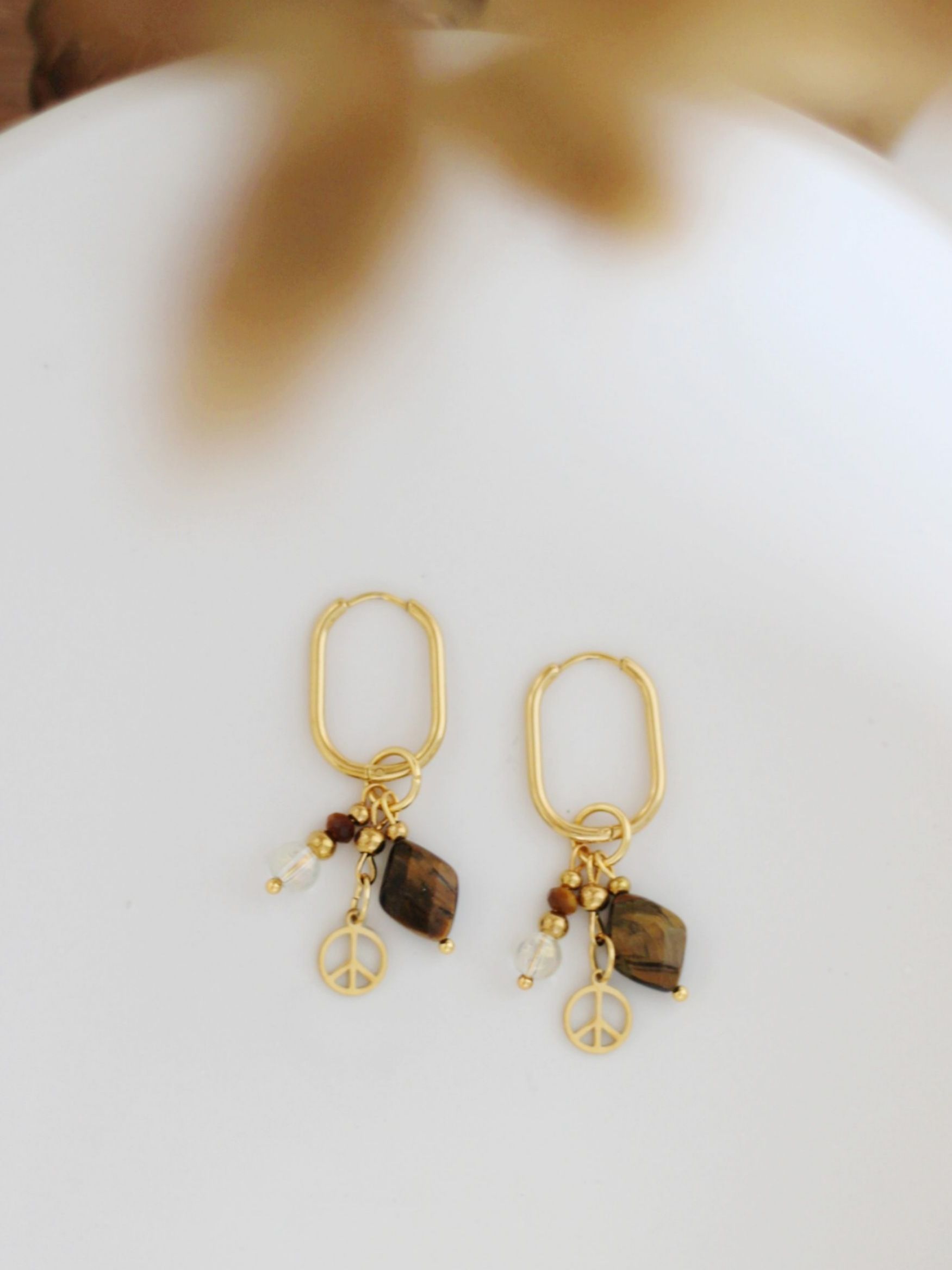 Ein Paar ovale Creolen in Gold mit langen Anhängern aus verschiedenen Perlen aus Tigerauge, Edelstahl und Peace-Zeichen