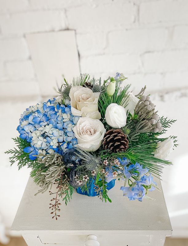 Close-up of blue hydrangeas, white roses, pinecones and evergreen greenery in a winter floral arrangement
105 characters