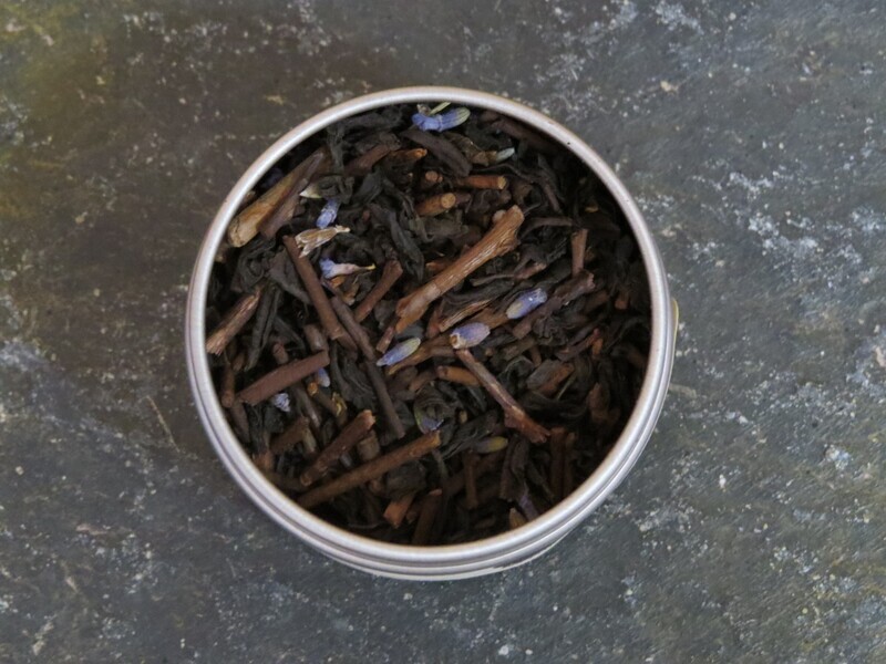 The image looks down into a small circular metal tin filled with a blend of green tea, black tea, and lavender. The tin sits on a textured green stone.
