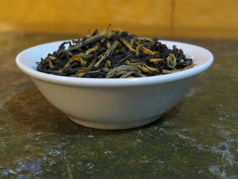 A small white bowl full of a blend of black and golden teas, slightly mounded, wood behind, stone underneath.