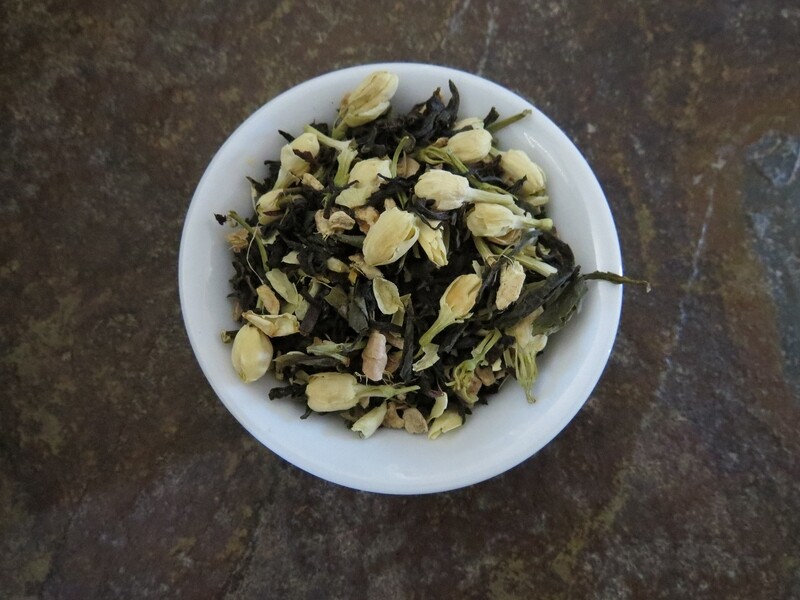 The image looks down at a small white bowl full of a blend of green tea, black tea, ginger, and jasmine.