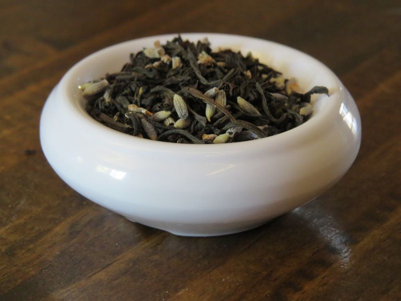 A small white bowl set on a dark grained wood, full of a blend of black teas and lavender.