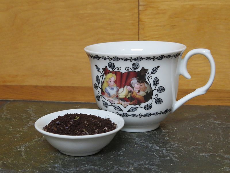 A small white bowl full of a blend of ground coffee and lavender flowers sits next to a cup that is shaped more for tea than coffee with a picture of Alice and the Mad Hatter prominent upon it.