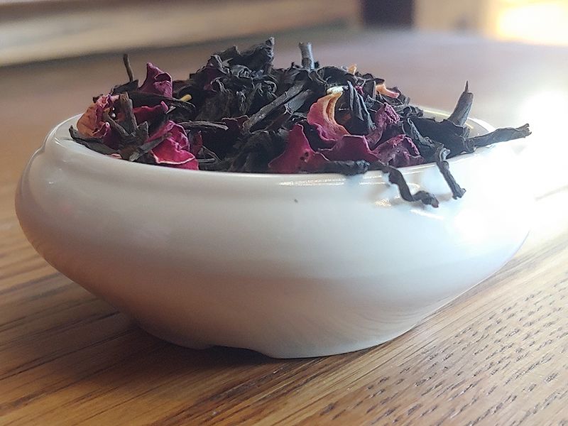 A small white bowl set on light and dark grained wood, lit from behind, so the edges of the bowl almost glow, drawing attention to the black tea drifting over the edge. Inside the bowl the tea is piled and blended with red rose petals and rosemary sprigs.