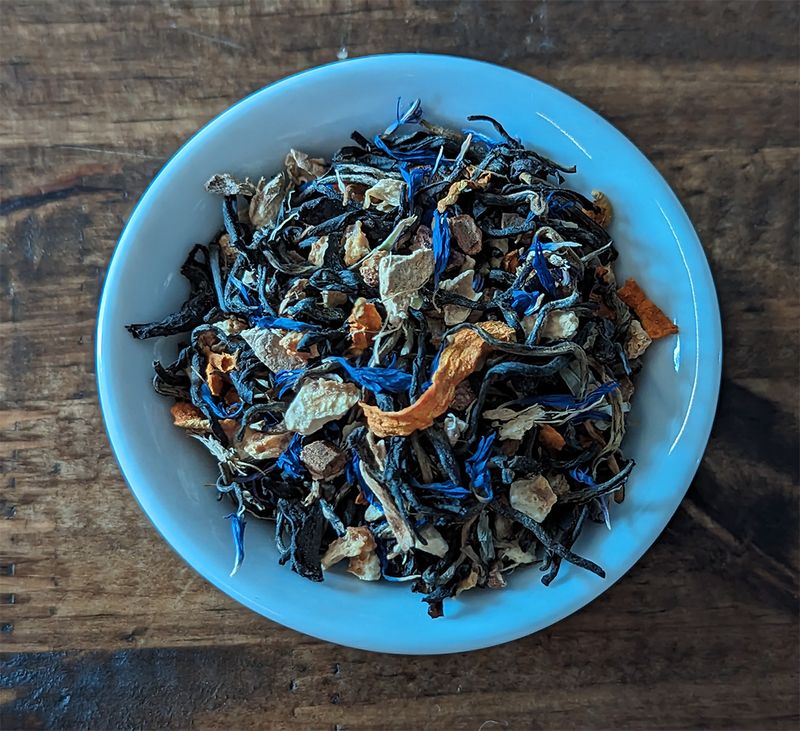 A small white bowl set on a dark wood background. The bowl is full of a blend of black teas, chais spices, vibrant orange turmeric curls, and bright blue cornflower petals.
