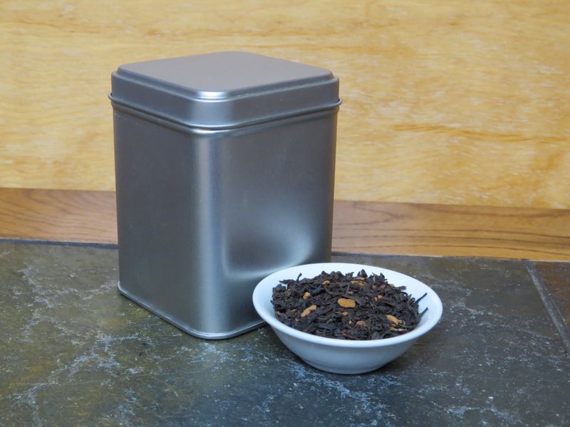 A small white bowl of black tea and cinnamon sits on a stone surface next to a silver tea tin.
