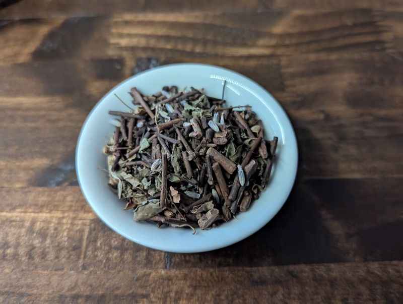 The image looks down at a small white bowl full of a blend of twig tea, a greenish purple herb, and lavender flowers.
