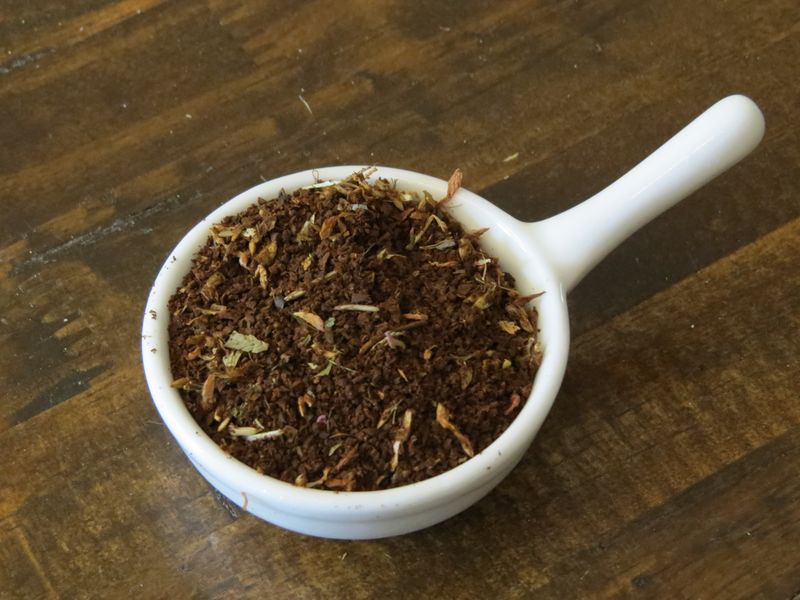 The image looks down into a small white handled bowl full of a blend of bits of fluffy red clover flowers and ground coffee.