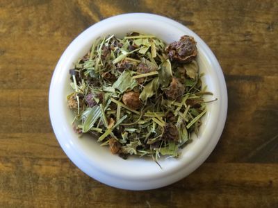 The image looks down into a small white bowl full of a blend of hawthorn berries, chicory, seaweed, and green herbs.