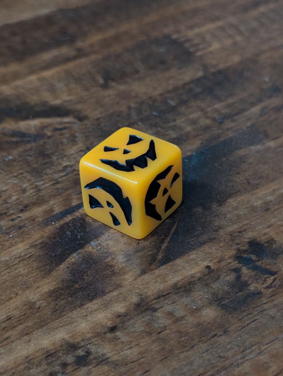 An orange and black d6, the numbers represented by pumpkin eyes and smiles.