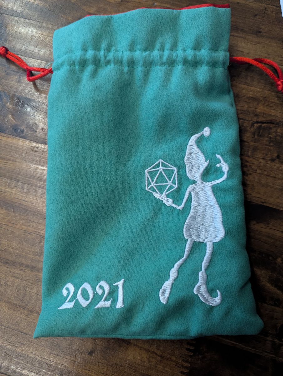 A dice bag with a soft green exterior, smooth red interior, red tie, with the image of a holiday elf holding a d20.