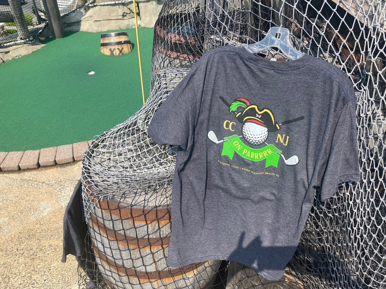 Dark grey t-shirt from Castaway Cove mini golf on Jenkinson&#39;s Boardwalk in Point Pleasant Beach. Back of shirt shown next to a hole at the miniature golf course. 